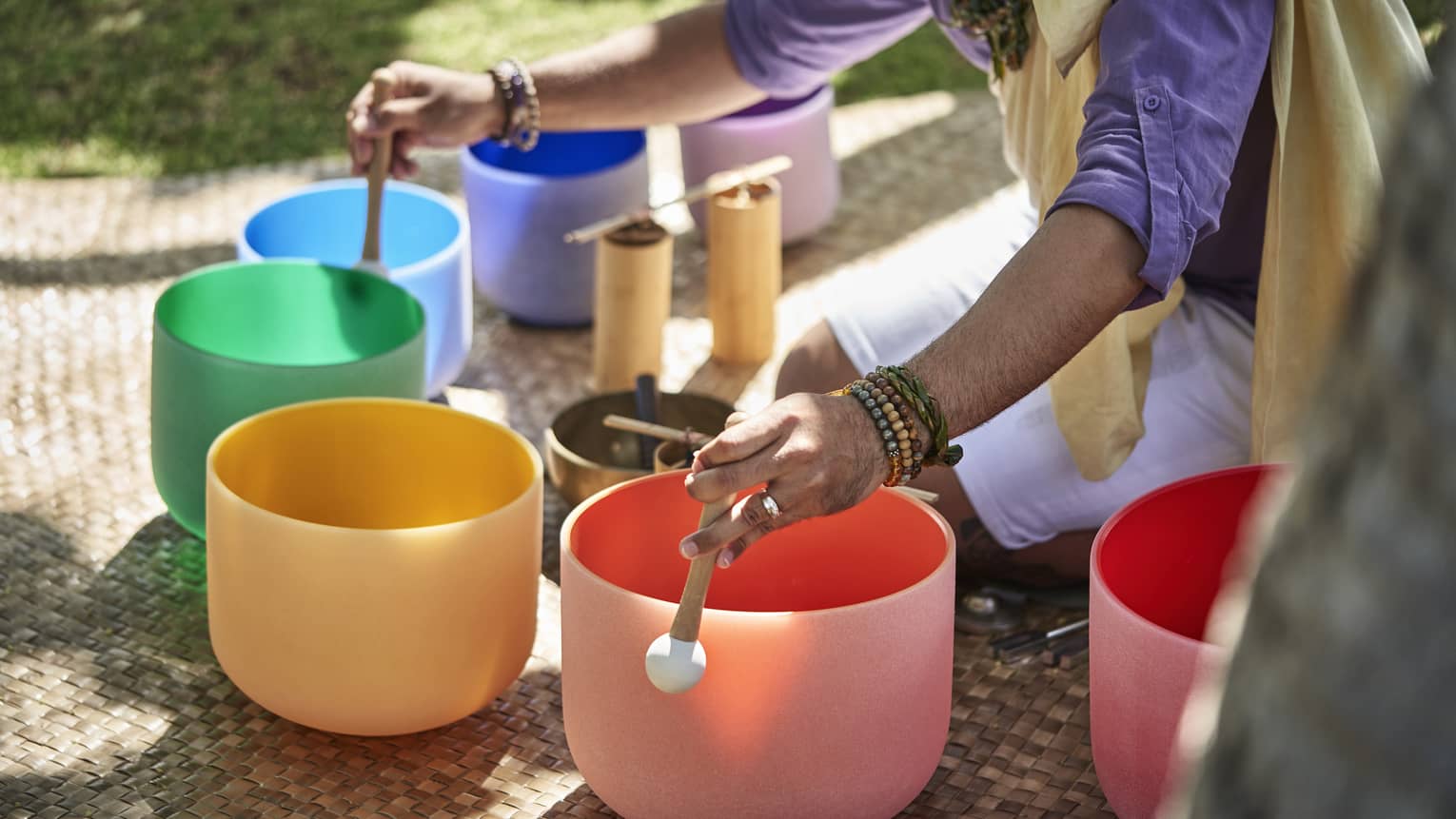Close up of a man's hands using mallets to play a row of colourful crystal sound bowls