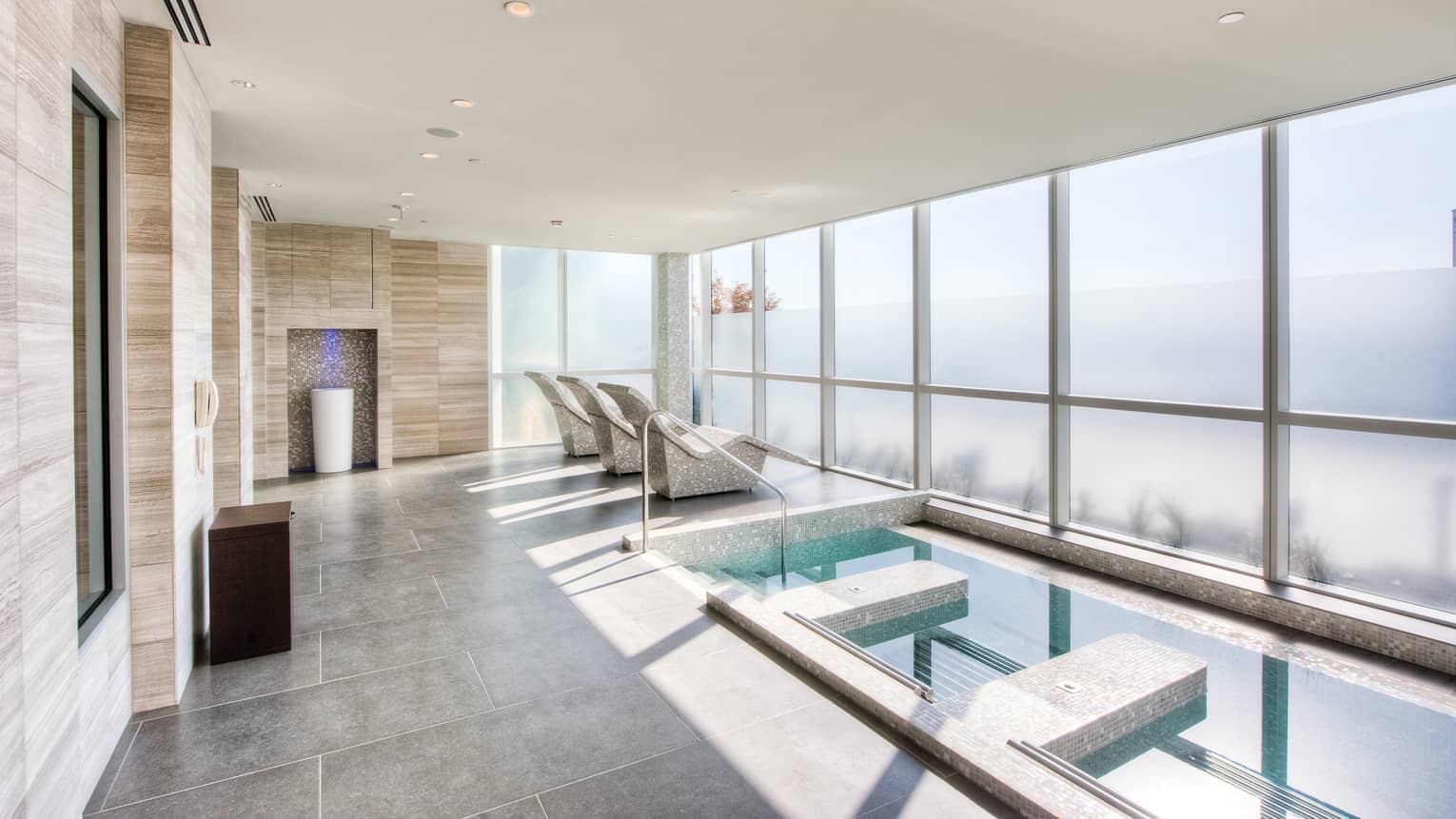 Spa plunge pool on grey tile deck, three stone chairs facing wall of windows 