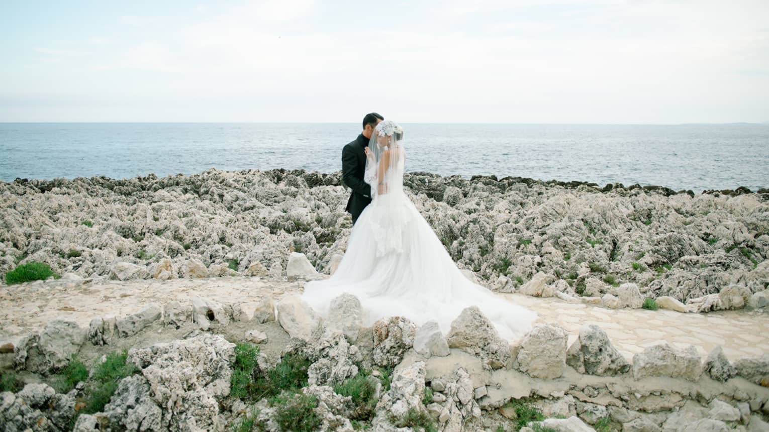 Groom and bride wearing veil over face in embrace, standing on rocky beach at sparkling blue sea