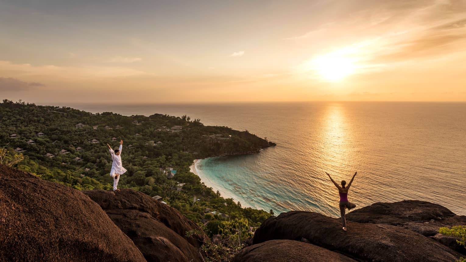 Woman and man stand apart on large boulders overlooking canopy of trees, ocean at sunrise 
