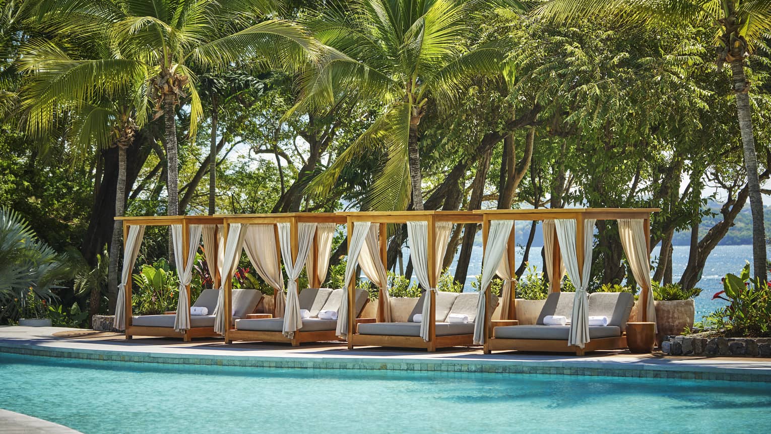 Four poolside cabanas sit next to a pool with a wall of tropical trees behind them
