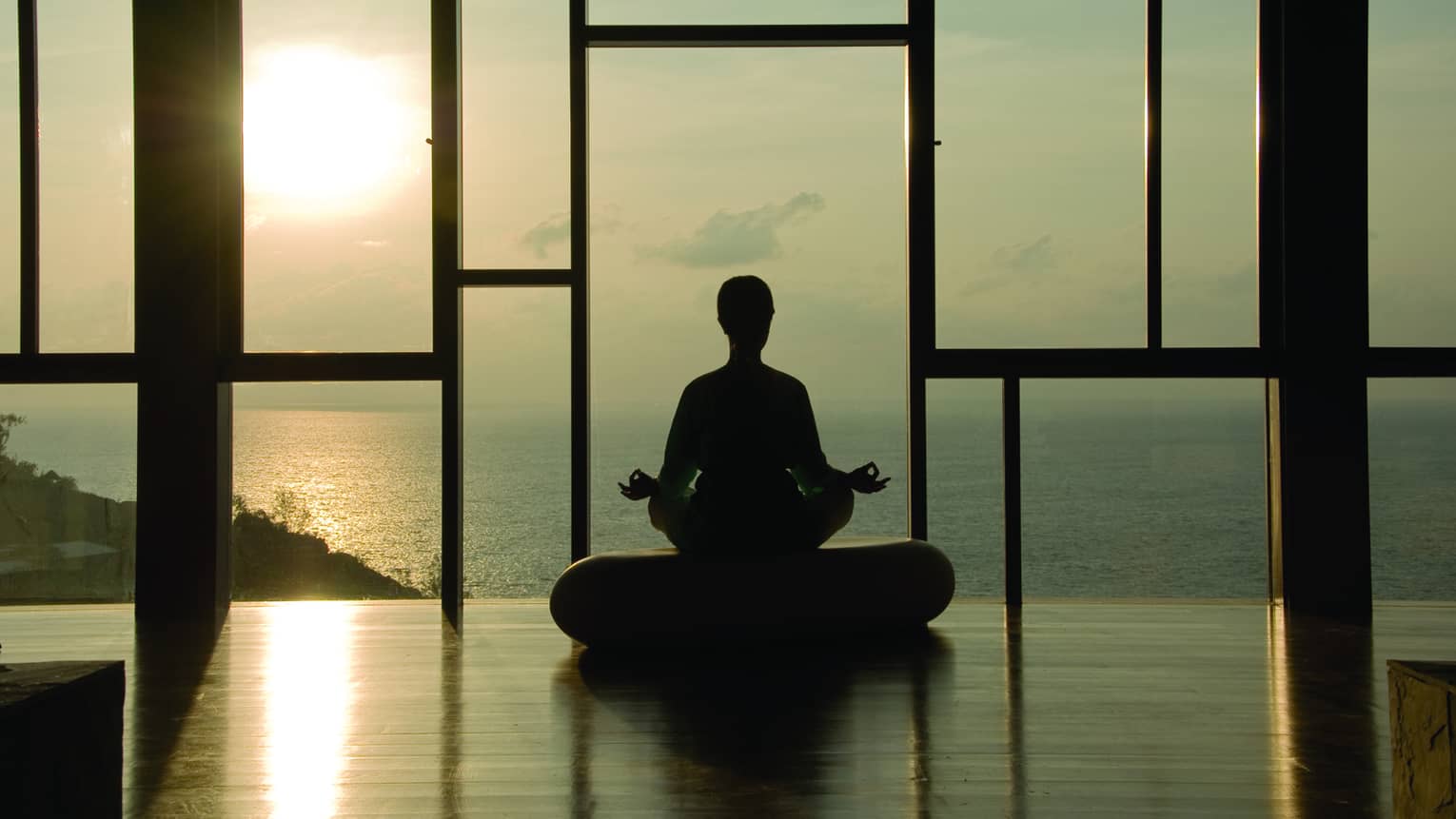 Silhouette of person meditating on cushion in front of floor-to-ceiling windows at sunset
