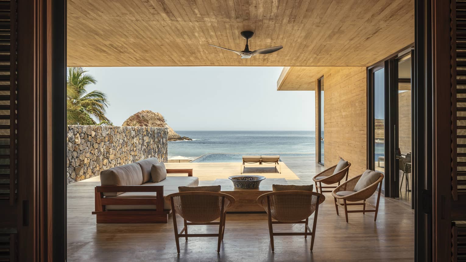 An open-air luxury villa overlooking the ocean, featuring a shaded seating area with modern wooden furniture and a private infinity pool extending towards the beach