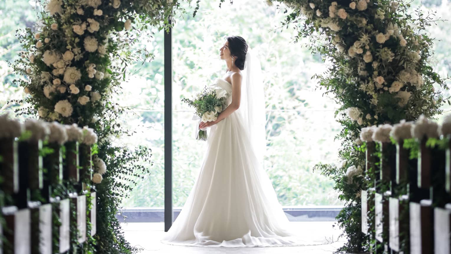 Bride stands under round wedding altar covered in white flowers by sunny window