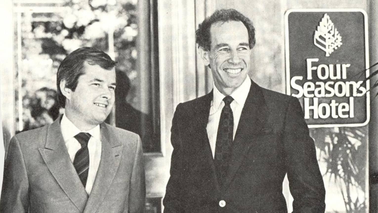 Black-and-white historic 1970s photo of two smiling men in suits by Four Seasons Hotel sign 