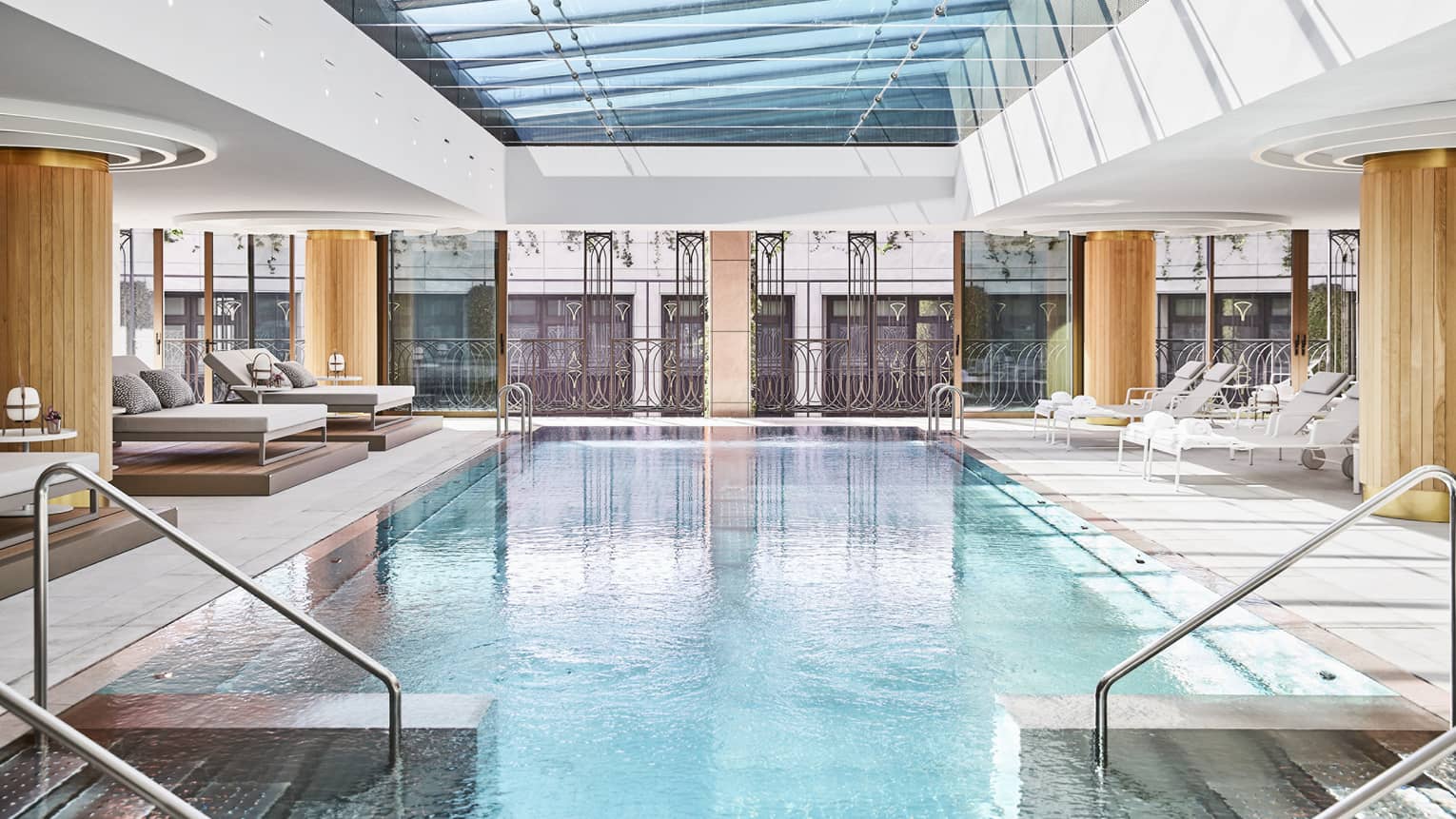 Luxury hotel indoor pool area with glass ceiling, metal railing accents and lounge chairs, surrounded by large windows and architectural detailing
