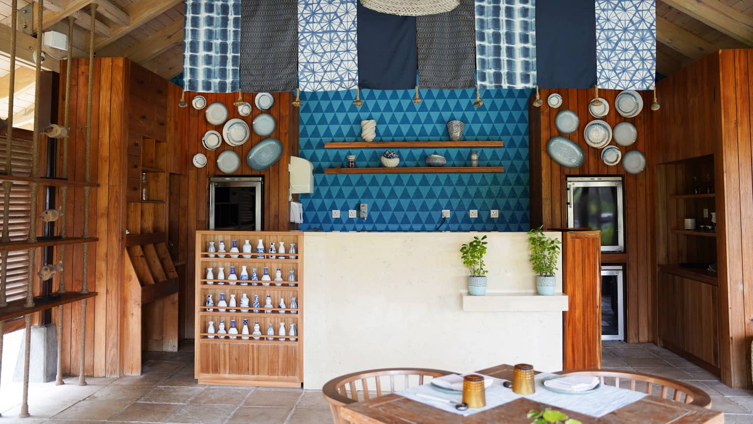 Casual restaurant with an island vibe, with blue and white fabric panels and rattan lights hanging from ceiling over teak and stucco bar