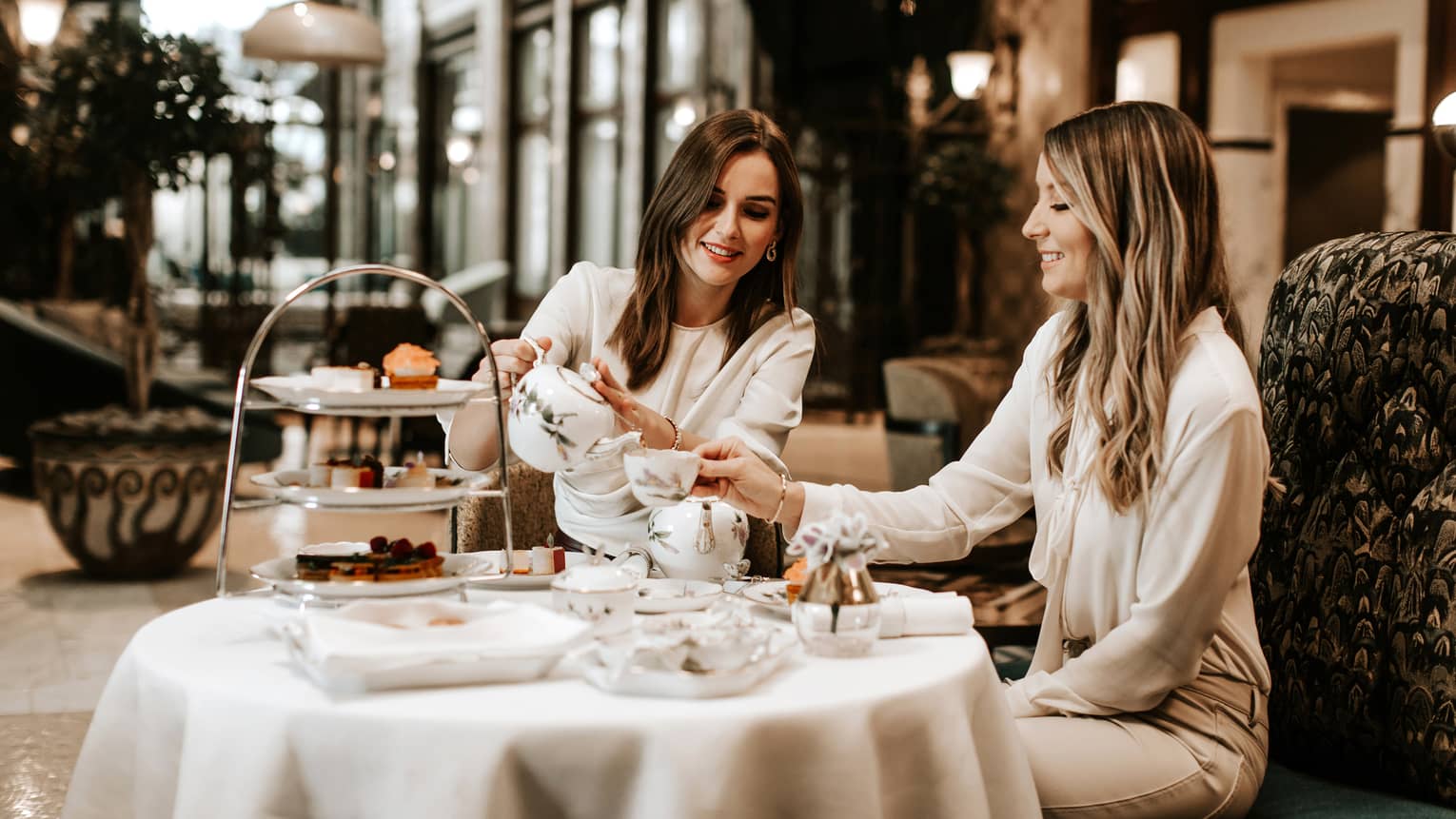 Two women in neutral outfits seated, smiling over afternoon tea in hotel lobby bar