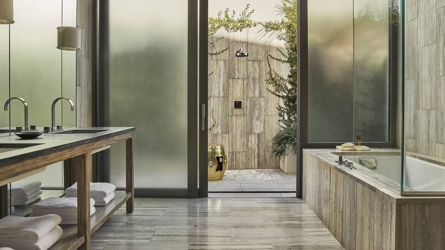 Modern bathroom with dual sinks, a soaking tub, frosted glass doors and an outdoor stone shower with greenery and a golden accent stool