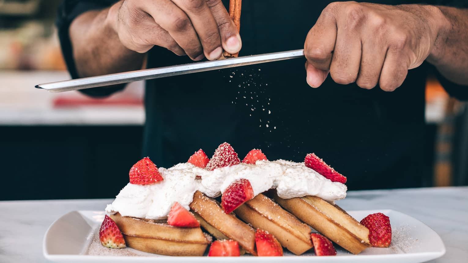 Close-up of waffles topped with whipped cream and strawberries, chef shaving cinnamon stick