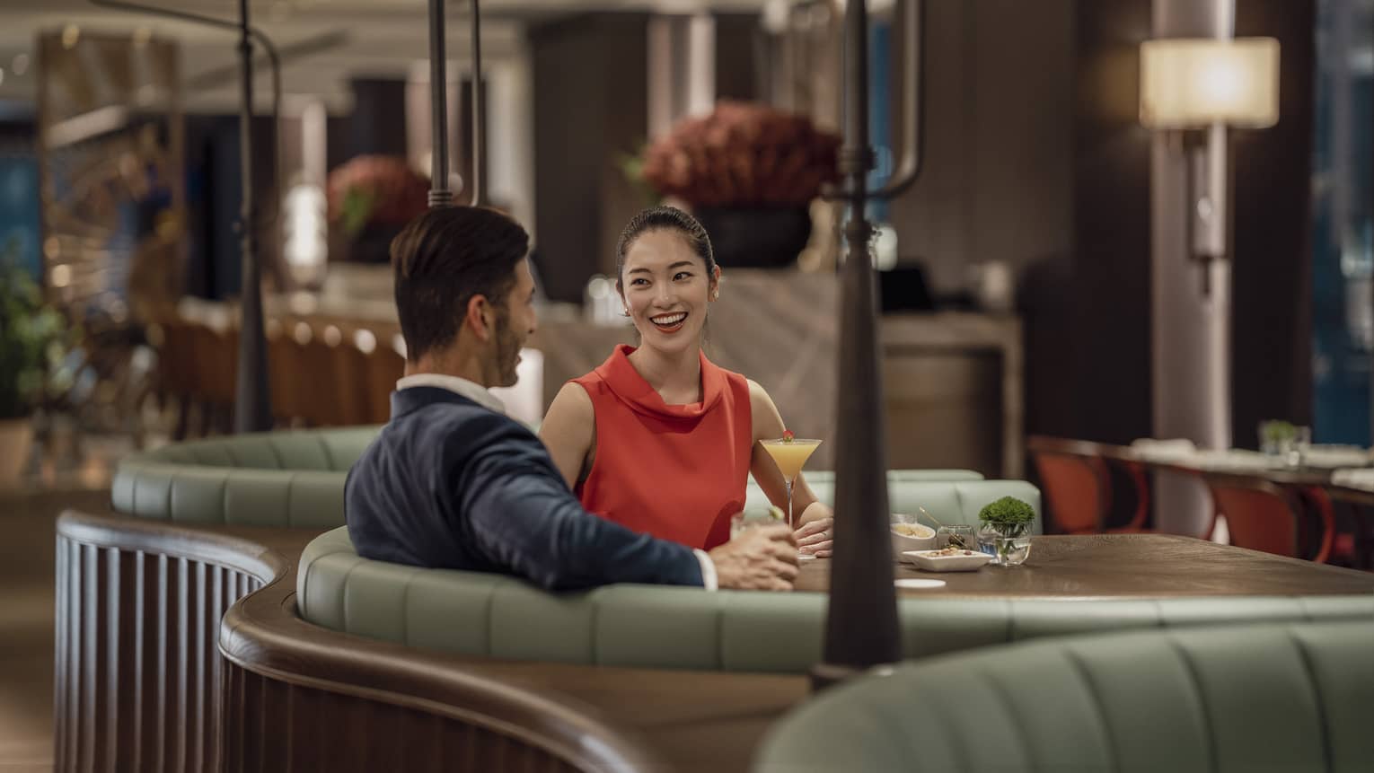 A smiling couple sit at a circular booth in an empty, dimly lit restaurant with cocktails and appetizers on their table.