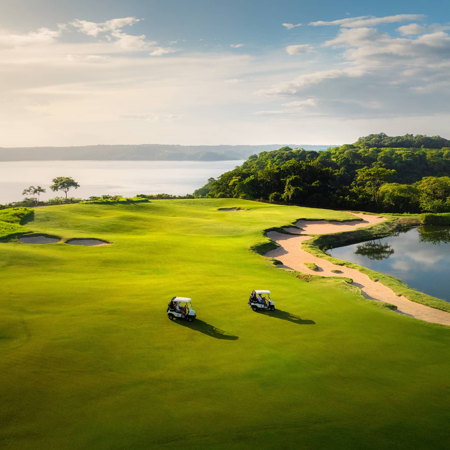Two golf carts ride along a green with the ocean in the background