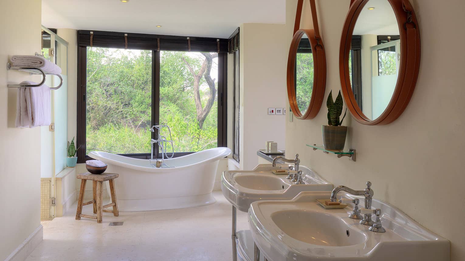 Bathroom with two white pedestal sinks against wall under two round mirrors and deep soaking tub in distance beside tall windows