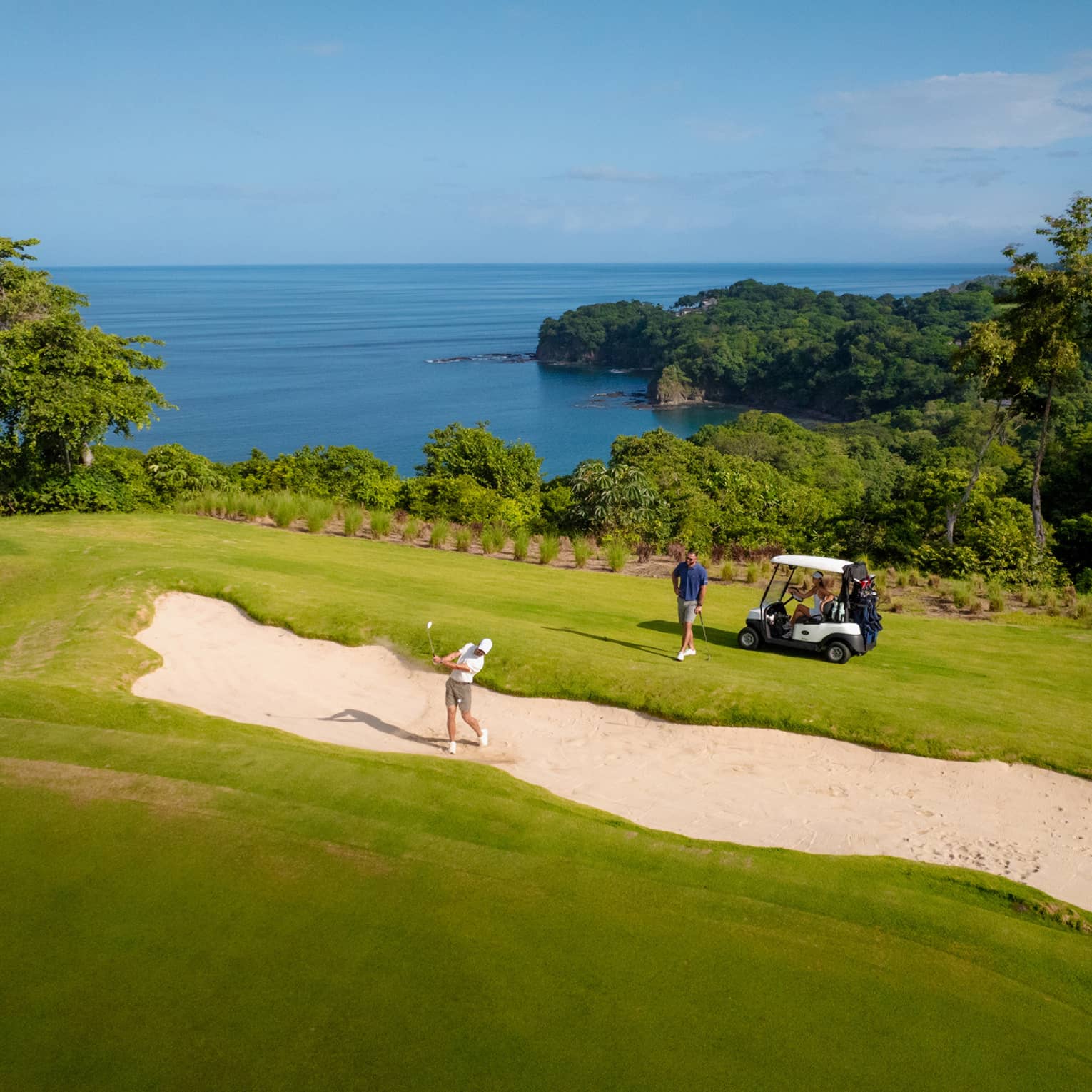 One golfer hits their ball out of a sand trap as another golfer stands by a golf cart looking on, with the ocean in the background