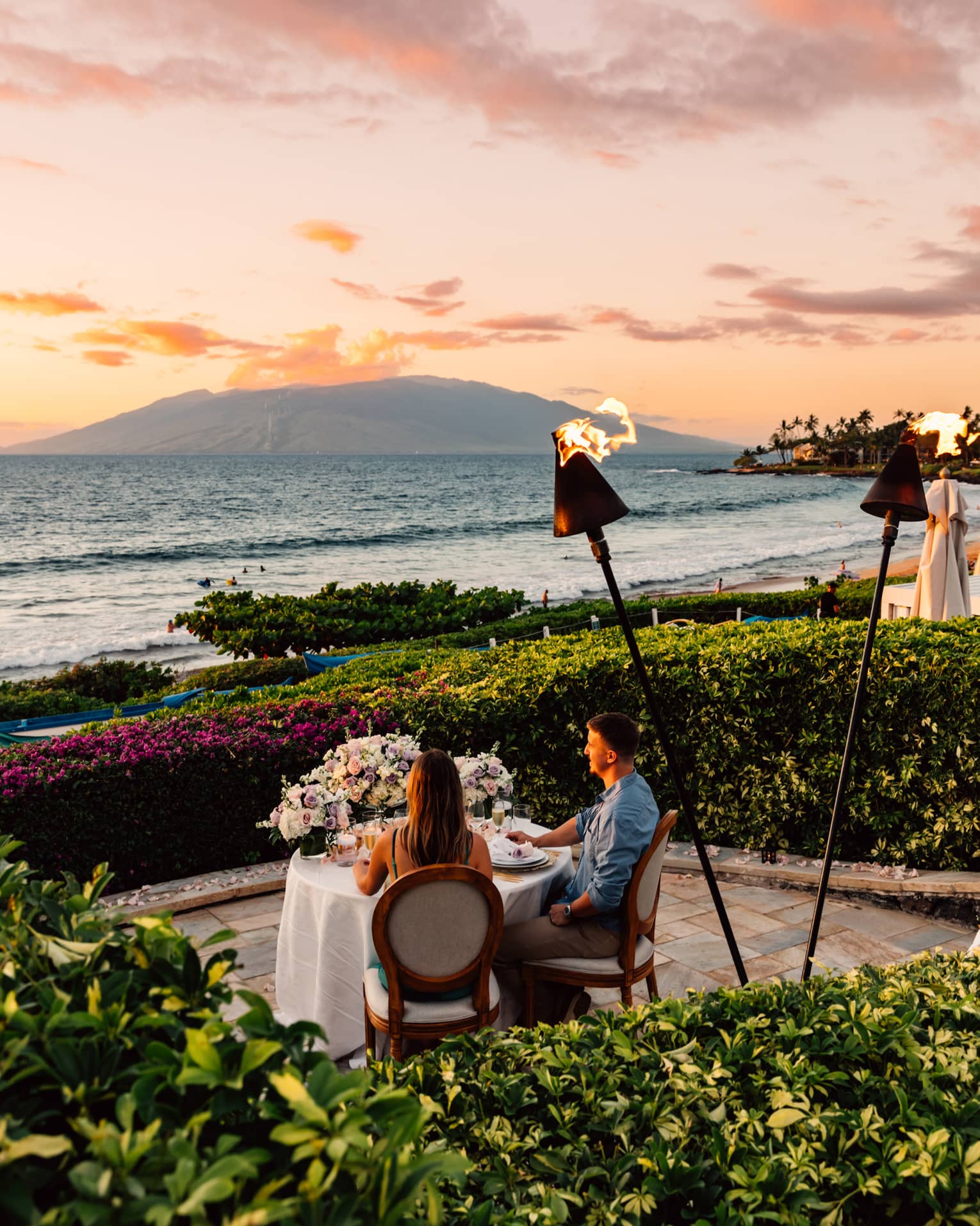 Couple dining at a table by the ocean at sunset with tiki torches and floral centerpiece