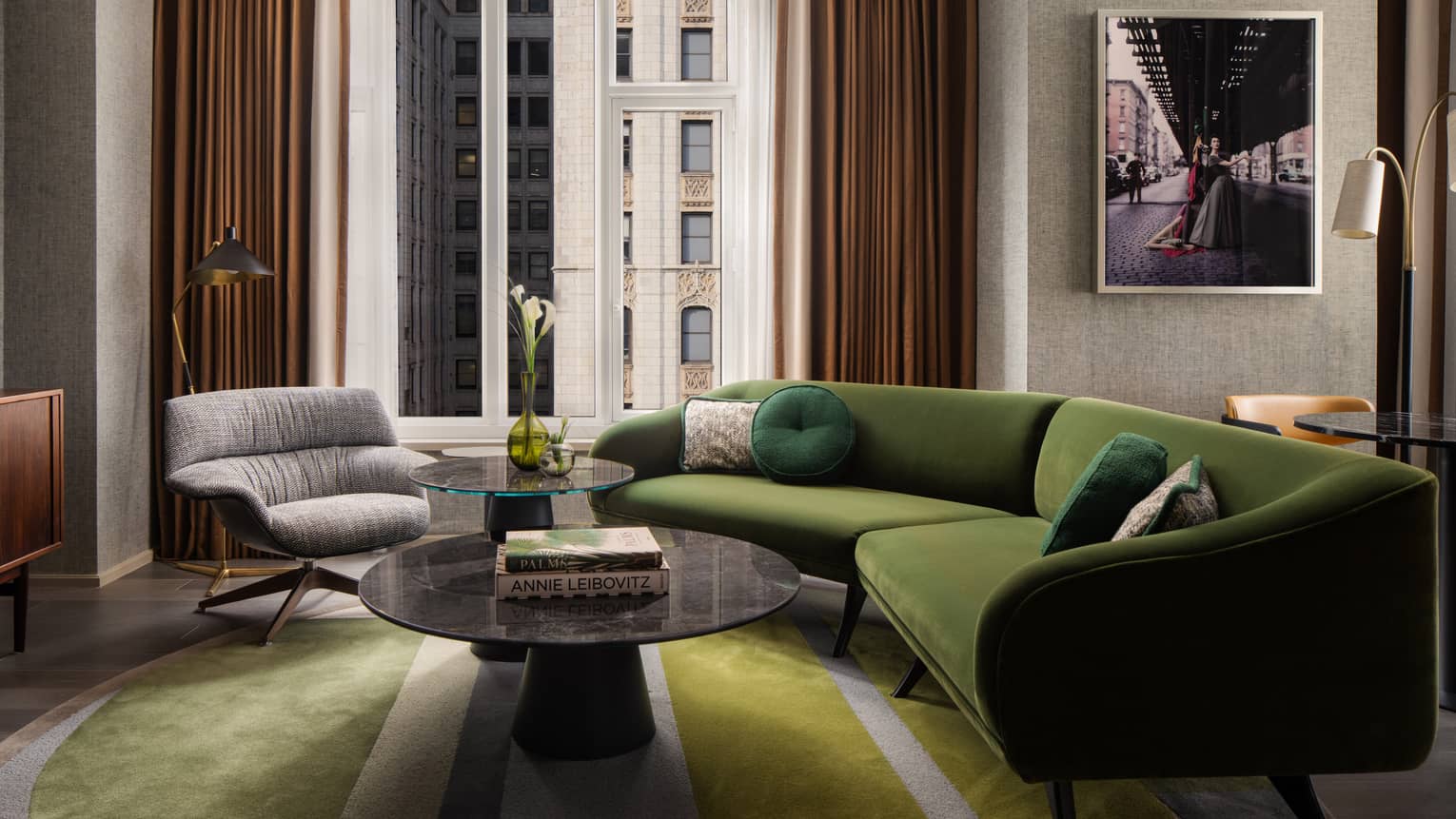 Suite living room with green sectional sofa, round coffee tables, gray armchair, wall art, and large window overlooking city buildings