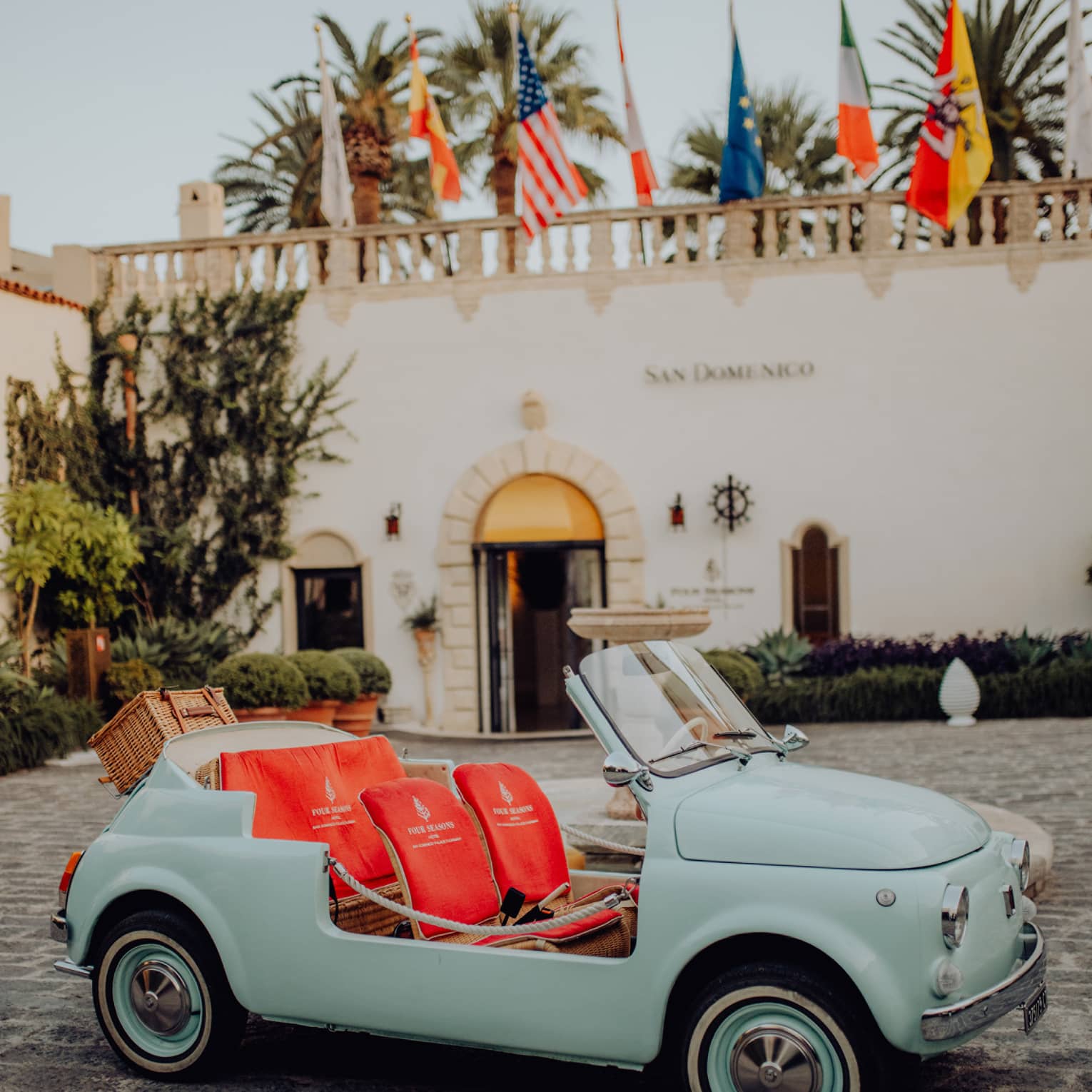 Vintage teal convertible Fiat 500 with bright coral seats parked on a cobblestone driveway in front of San Domenico Palace.