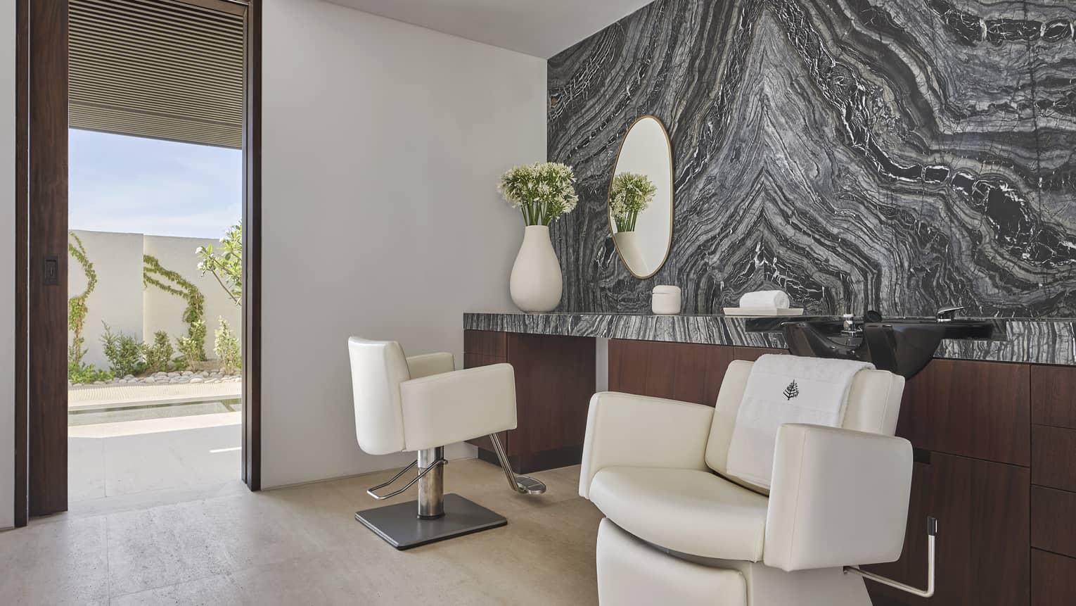 two chairs in a room with a large shelf and grey wall, a door looking at plants outside is open.