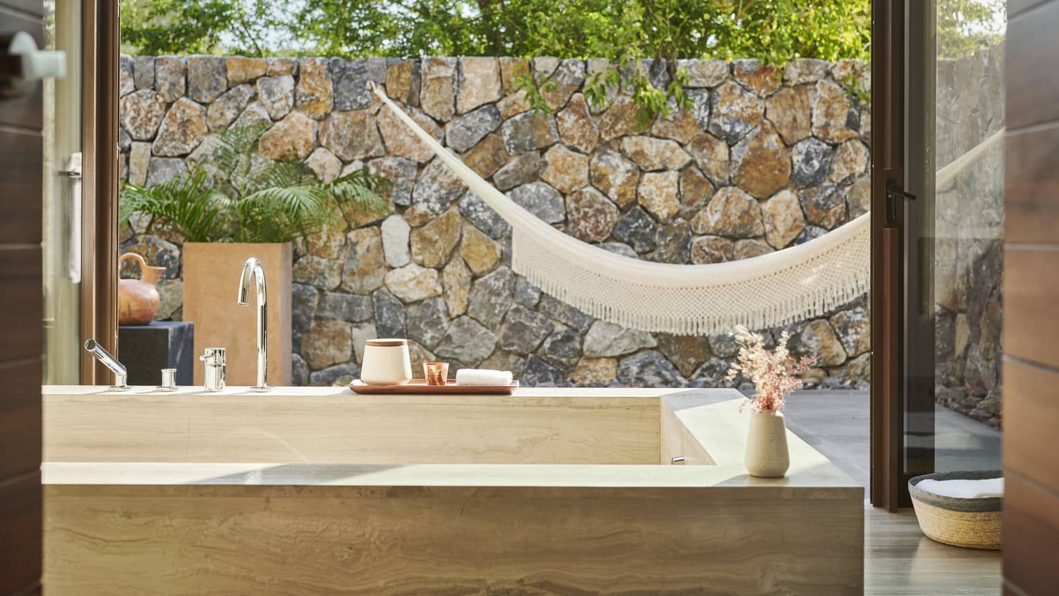 A bathroom with a large bath and large window looking outside at a hammock and stone wall.