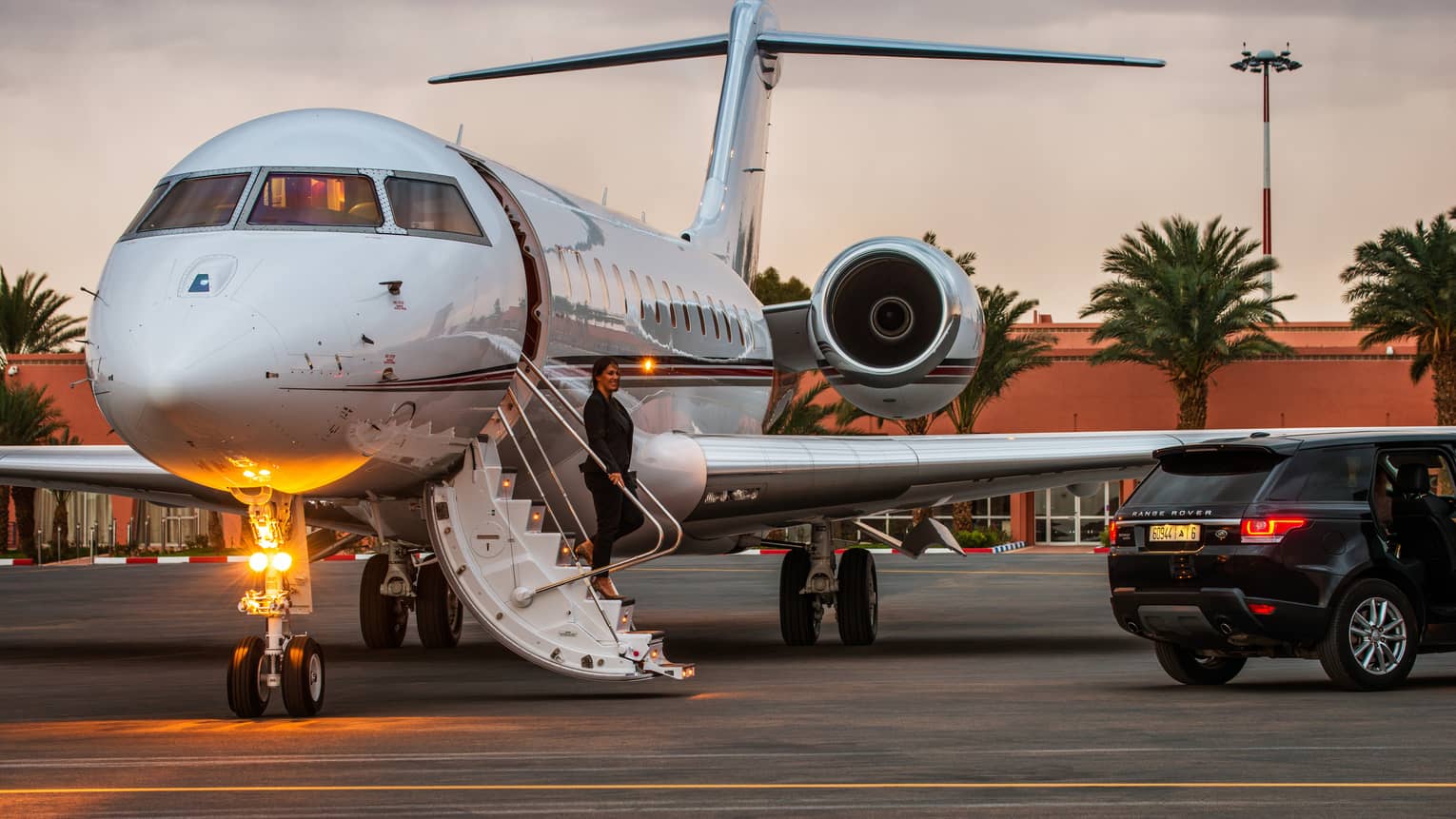 A woman stands on stairs of private plane, a black SUV is parked just outside