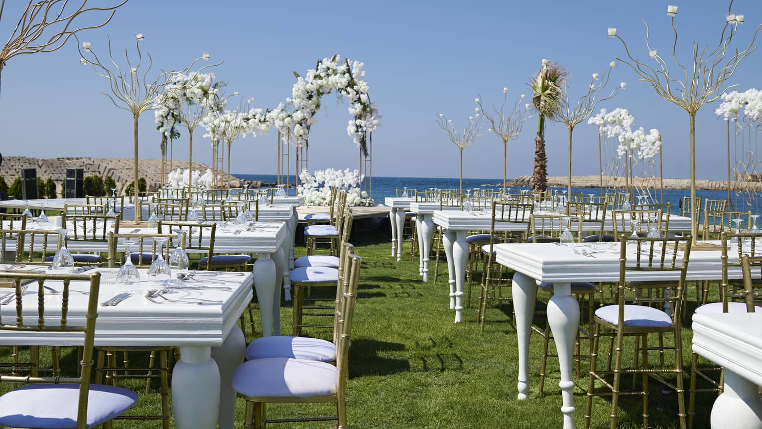 Oceanfront lawn set for a wedding with white tables, gold chairs and white orchids