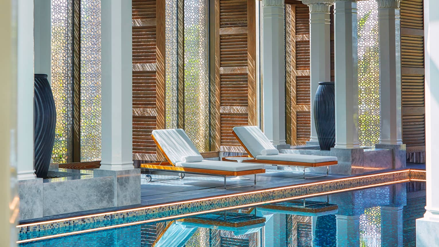 Two white lounge chairs on deck beside white pillars, in front of indoor swimming pool