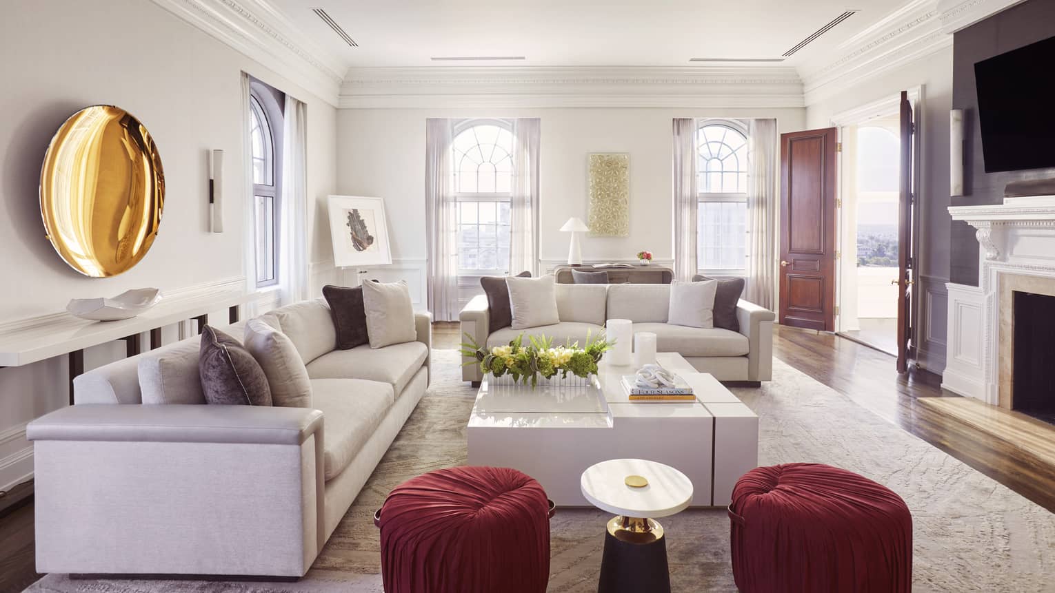 Living room with two sofas, large white square coffee table, two red cushioned stools, gold plate artwork on wall, TV and fireplace
