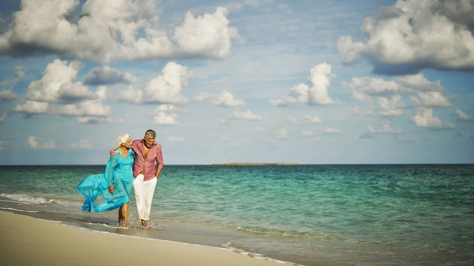Two people walking along a tropical beach with turquoise waters under a partly cloudy sky