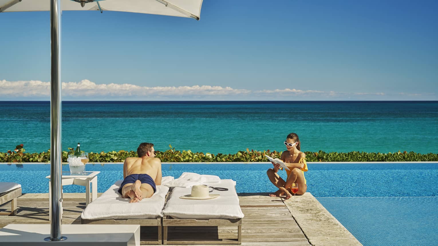 Person relaxing on a sun lounger under an umbrella, while another person reads by the infinity pool overlooking the ocean