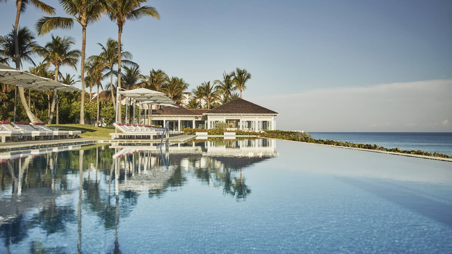 Blue sky, palm trees reflected on large ocean pool, bungalow in background