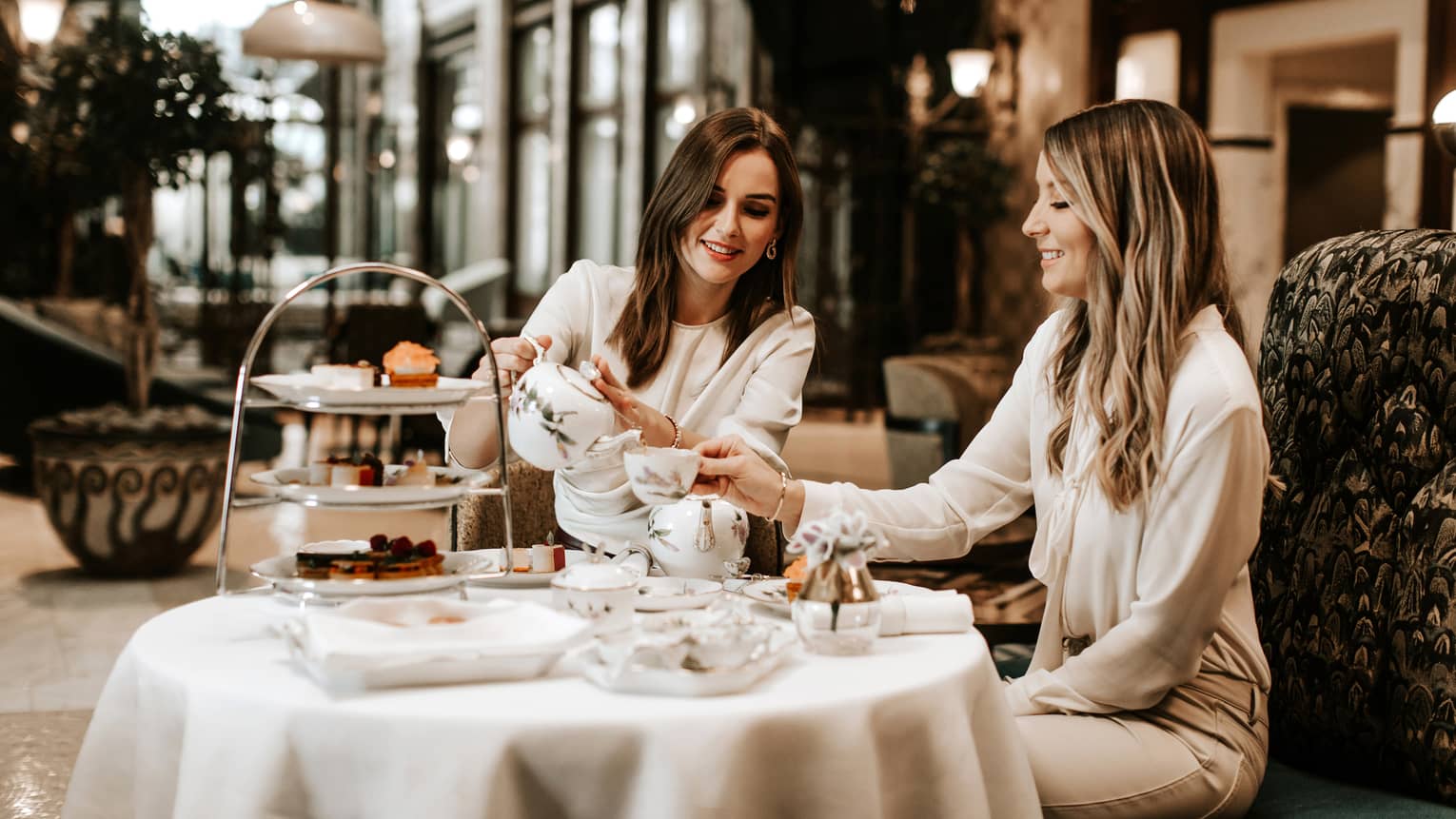 Two women in neutral outfits seated, smiling over afternoon tea in hotel lobby bar