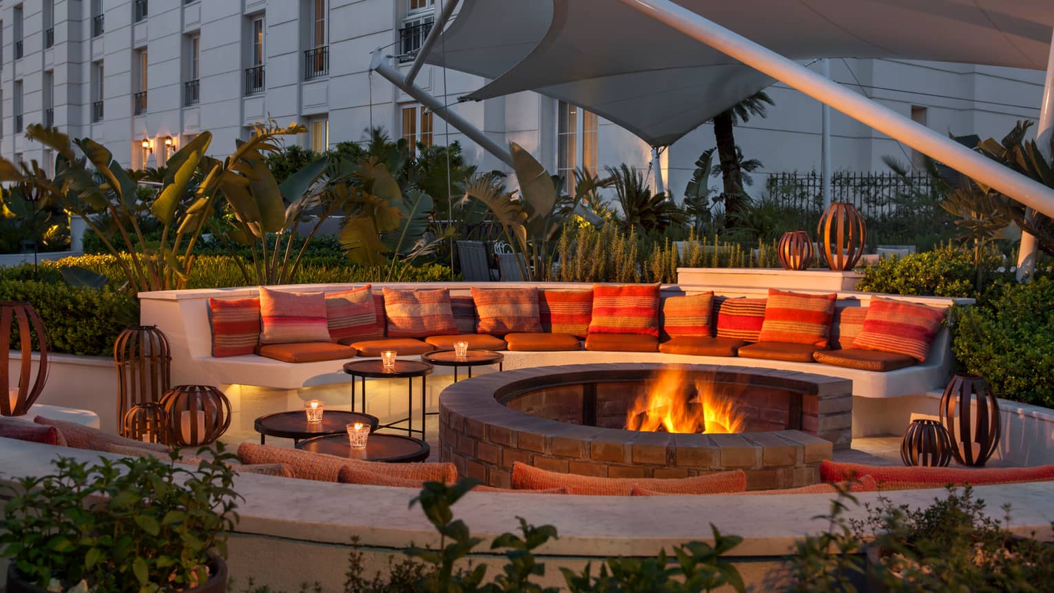 Large curved white bench with cushions around outdoor fire pit with flames at dusk
