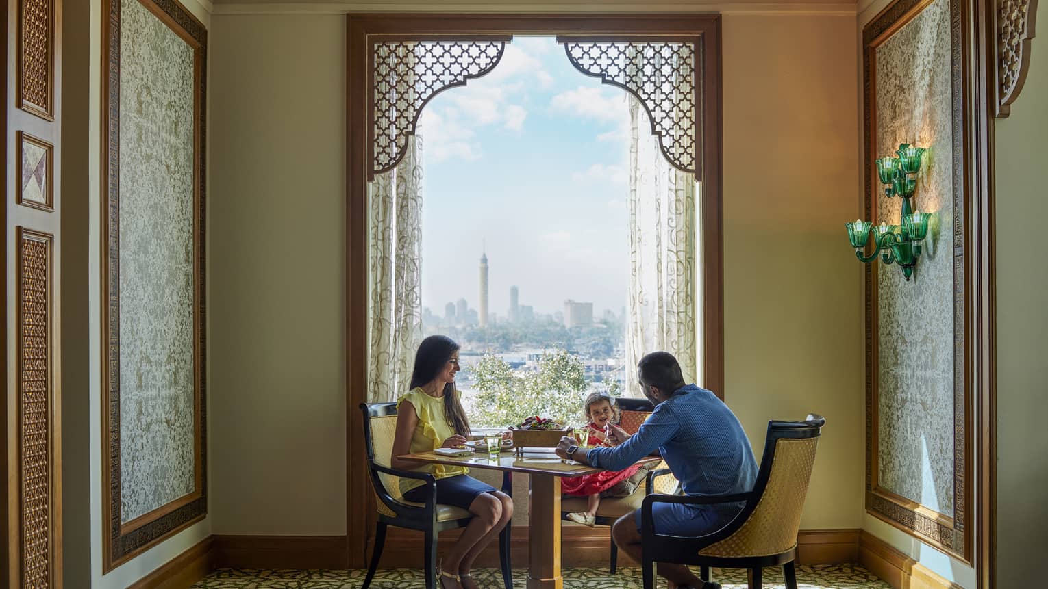 Family of three seated at dining table in front of tall window with city views
