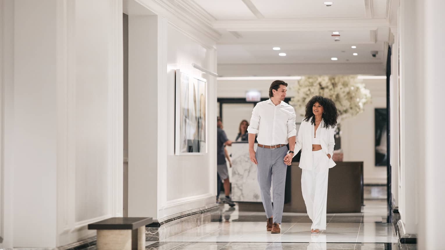 A man and woman walking down a hotel hallway with a lobby behind them.