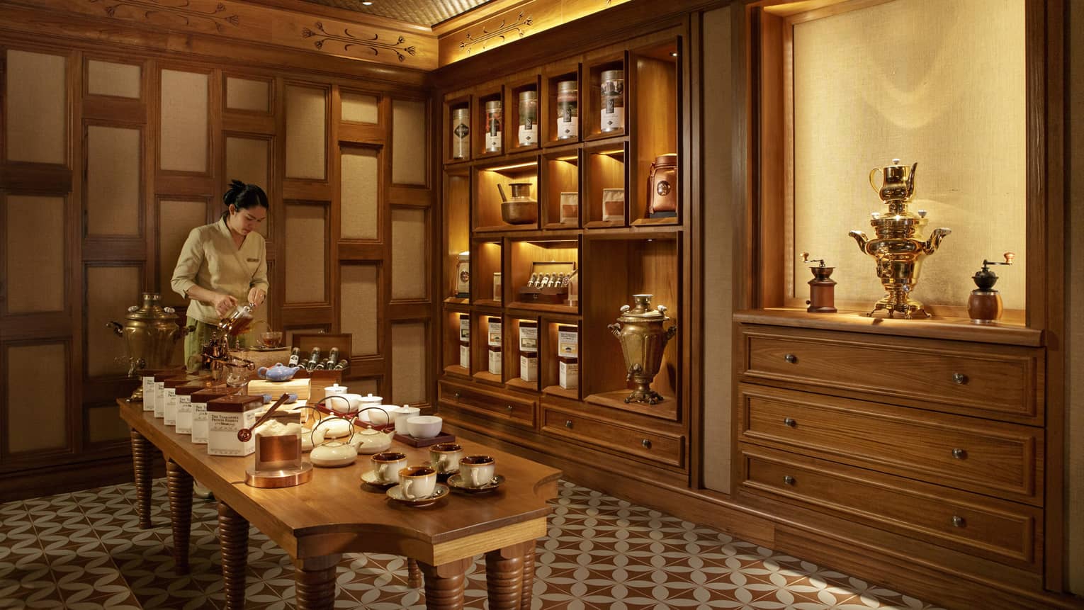 Server prepares tea at long table with pots, cups in front of wood panel wall, pantry
