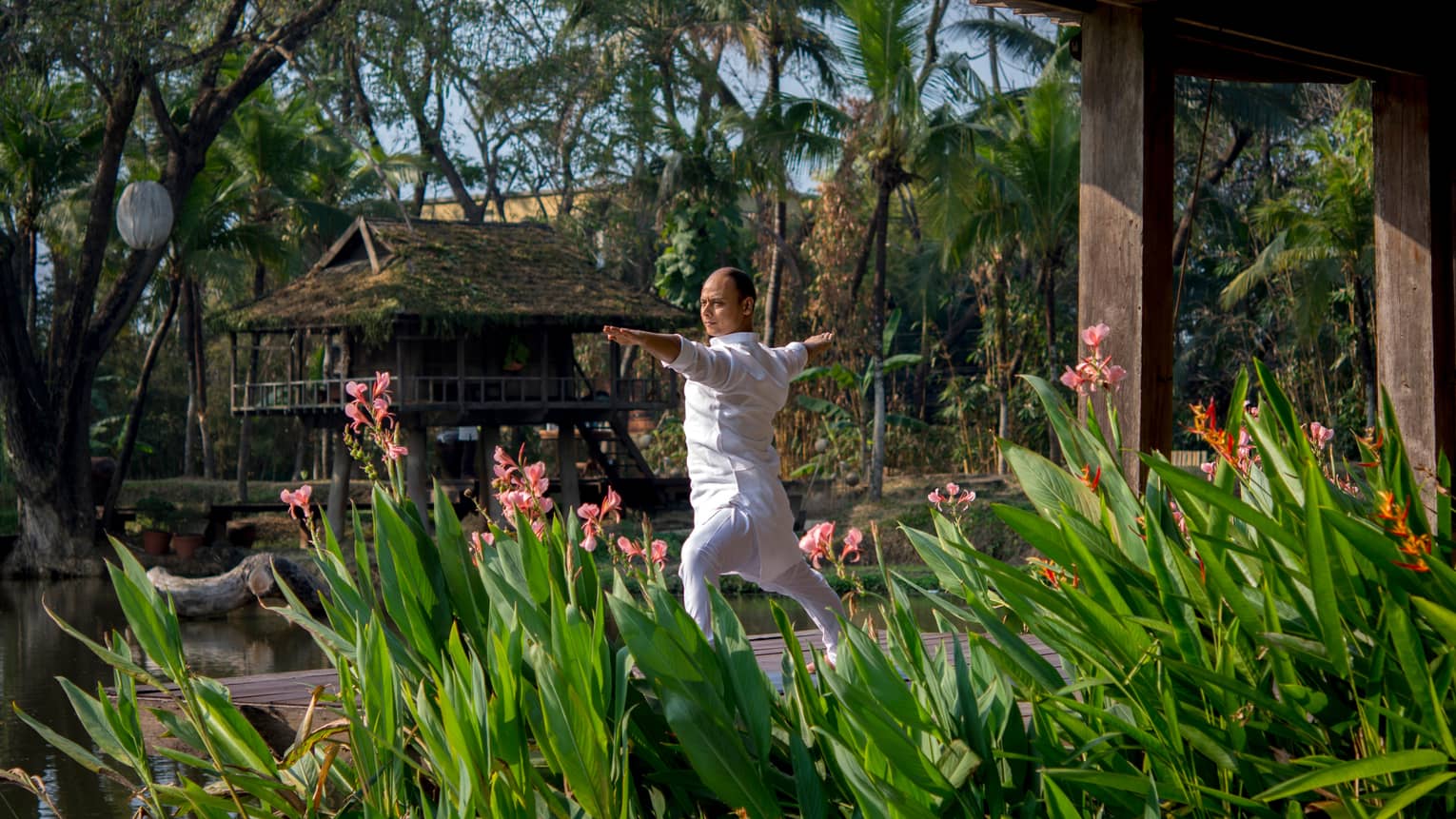 Resident yogi Dheera wearing white in yoga pose with arms outstretched by tall flowers, leaves
