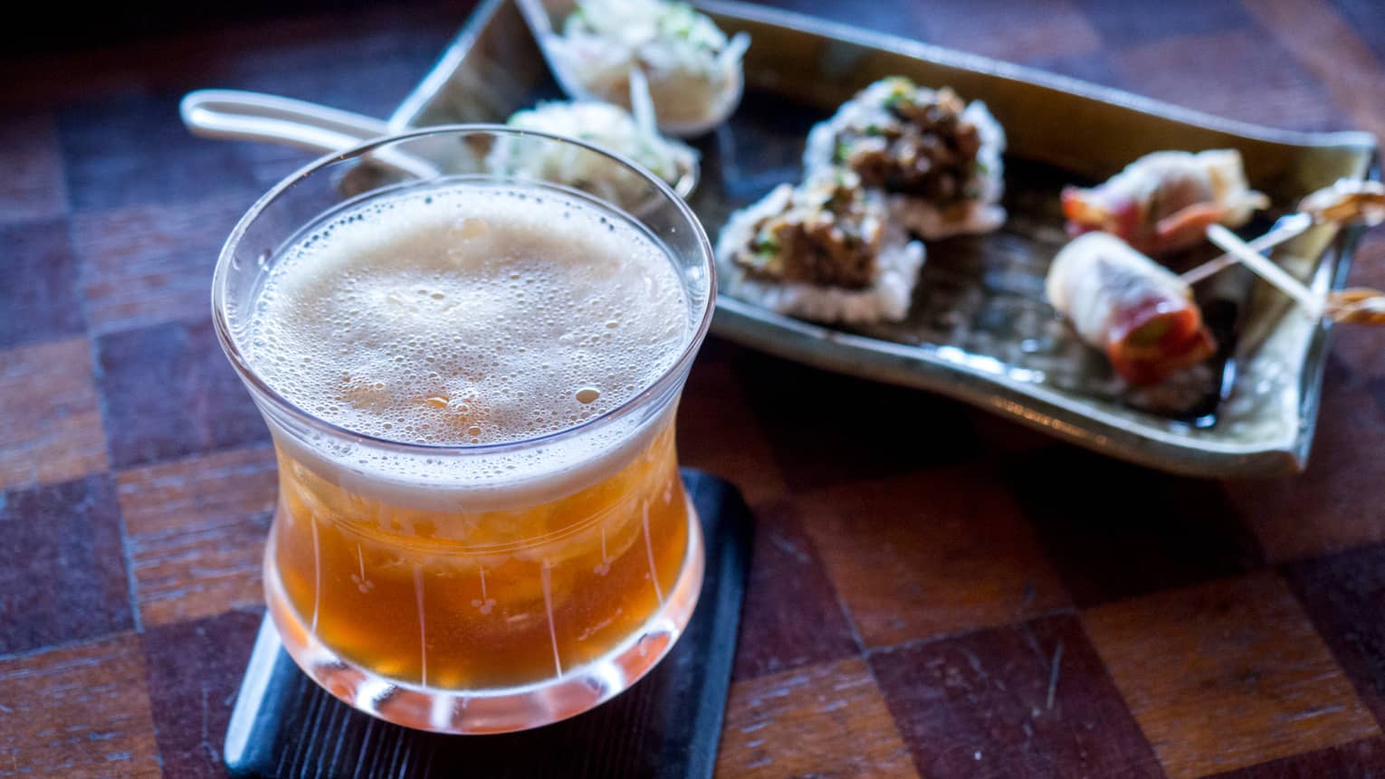 Frothy cocktail in rock glass beside platter with small appetizers 