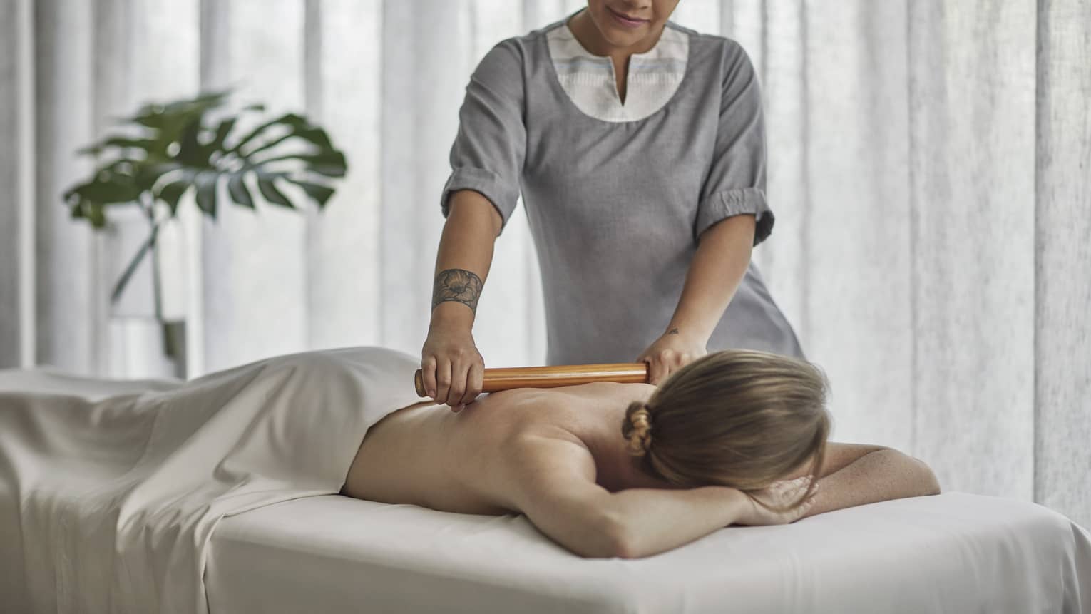 Spa therapist wearing a grey tunic rolls a wooden roller on the bare back of a guest who is laying on a massage table