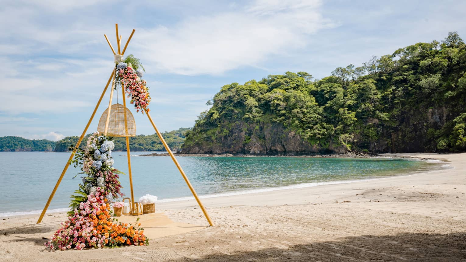 Four wooden posts strung together in a pyramid shape with various flowers strung around it sits on a beach by the water