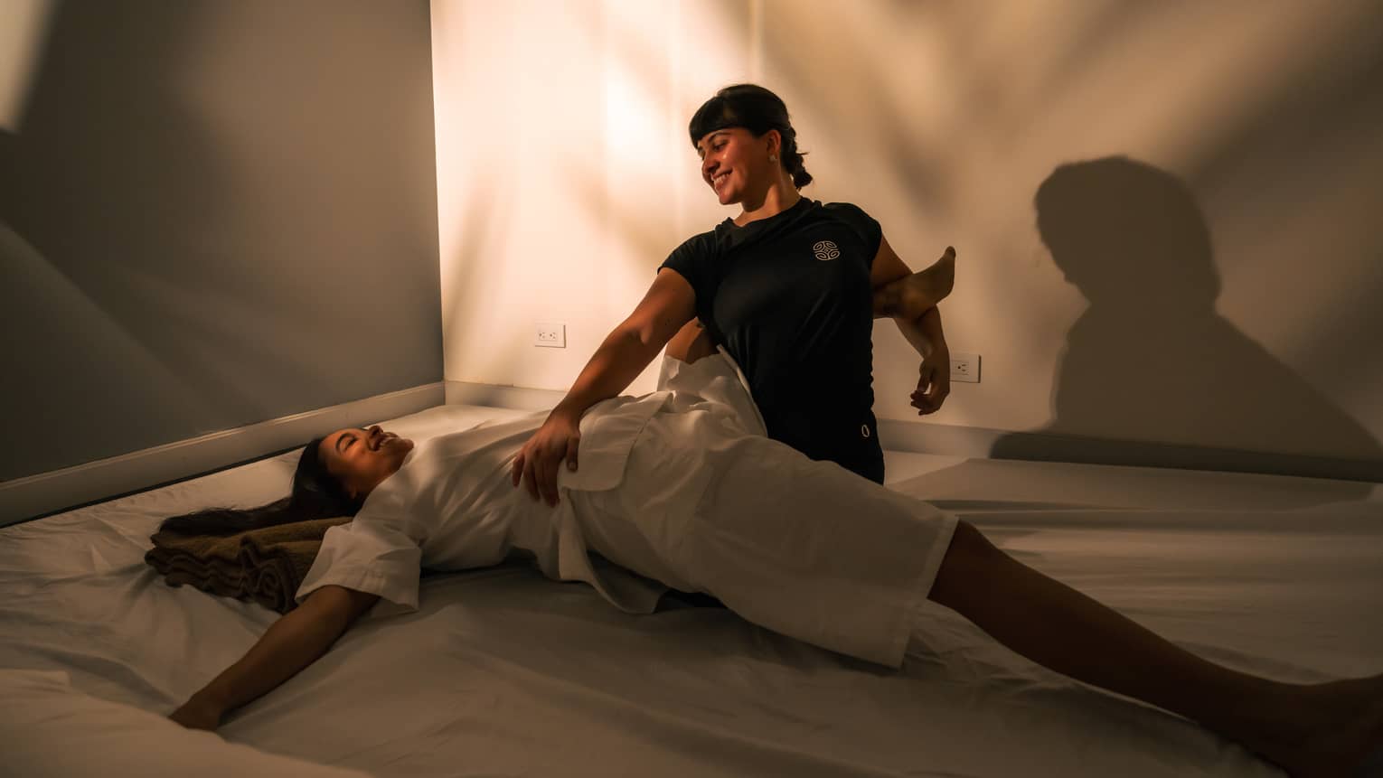 Two people sit in shadowy room as one of them helps stretch the back of the other
