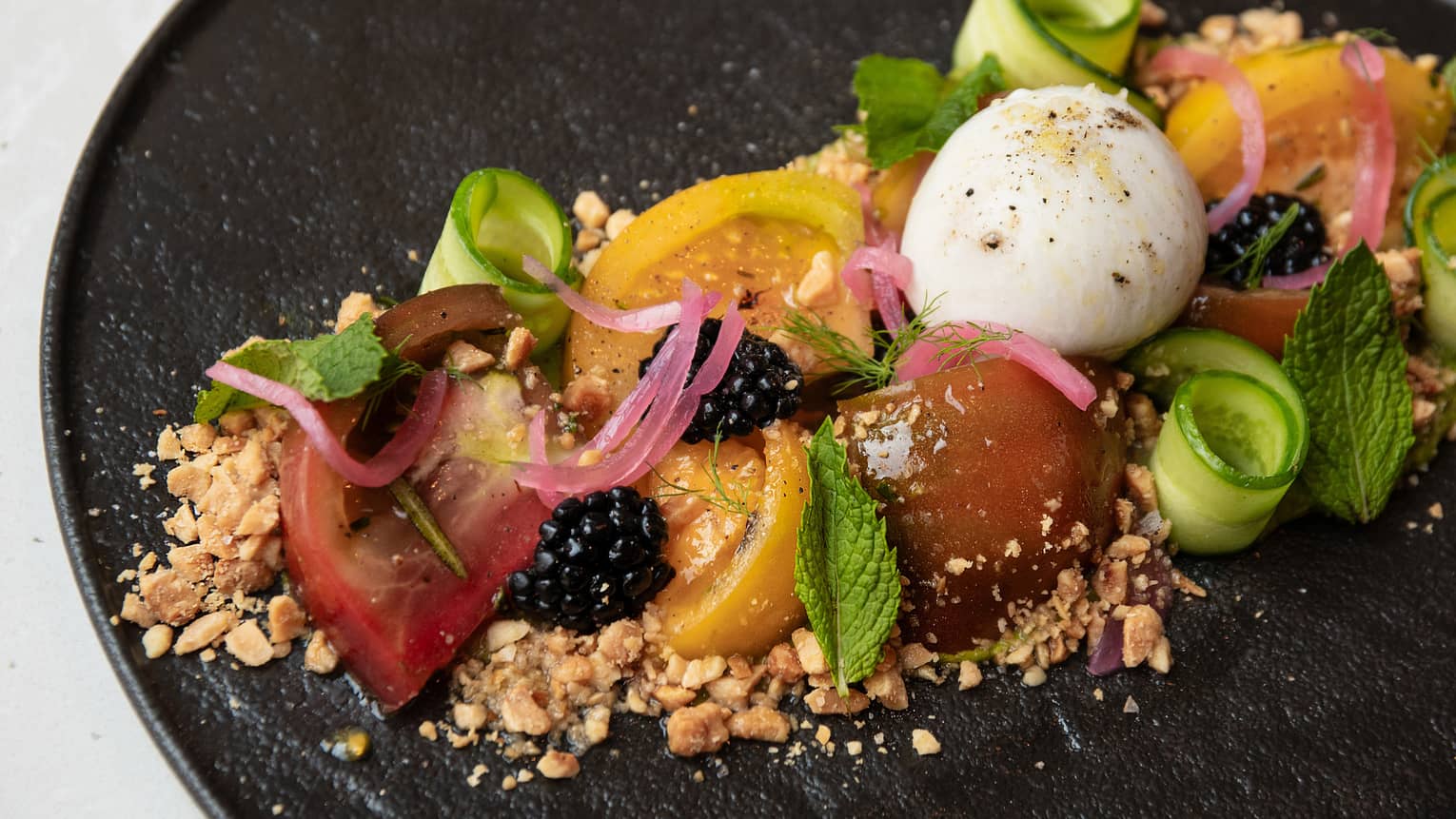 Heirloom tomato salad with crushed almonds, rolled cucumber slices, blackberries, pickled shallot and fresh mint plated on a round, black dish