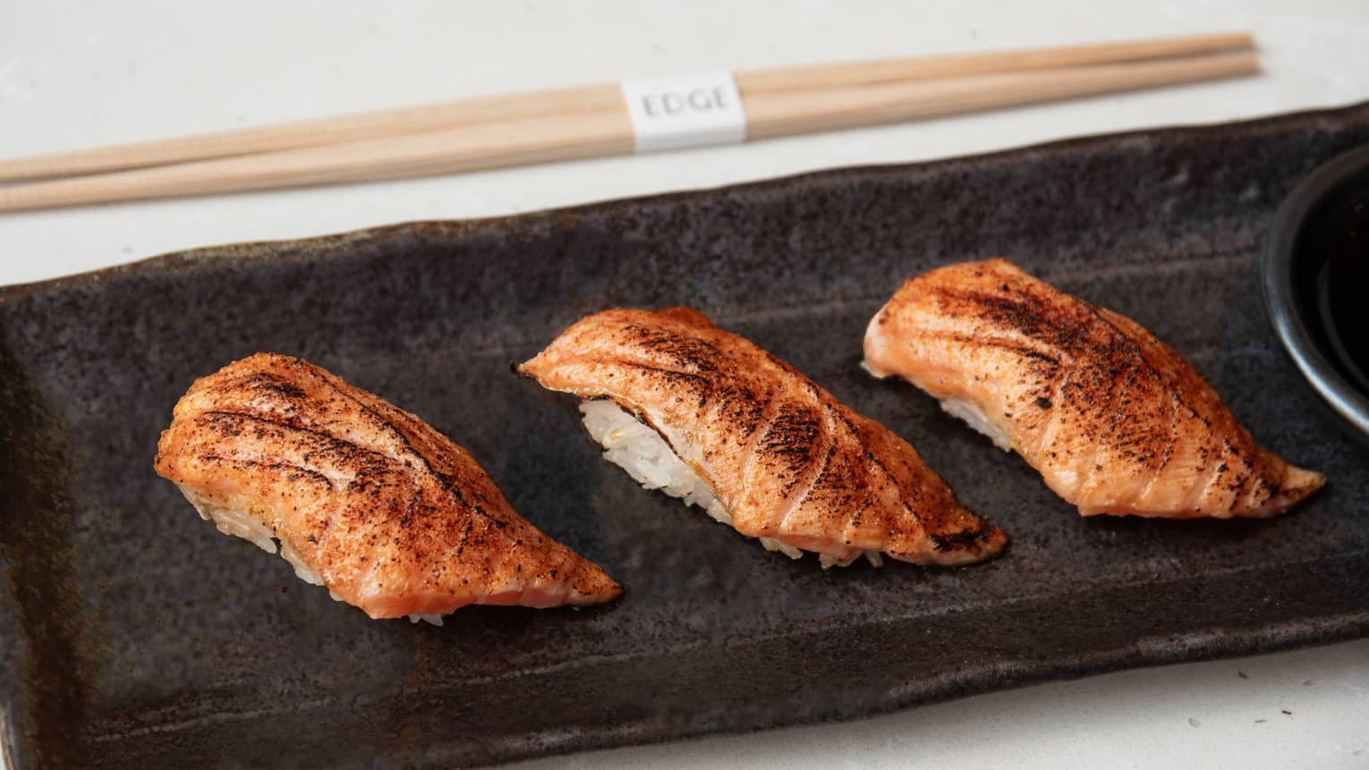 Three pieces of sorched salmon nigiri in a row on a black rectangular plate with chopsticks to the side
