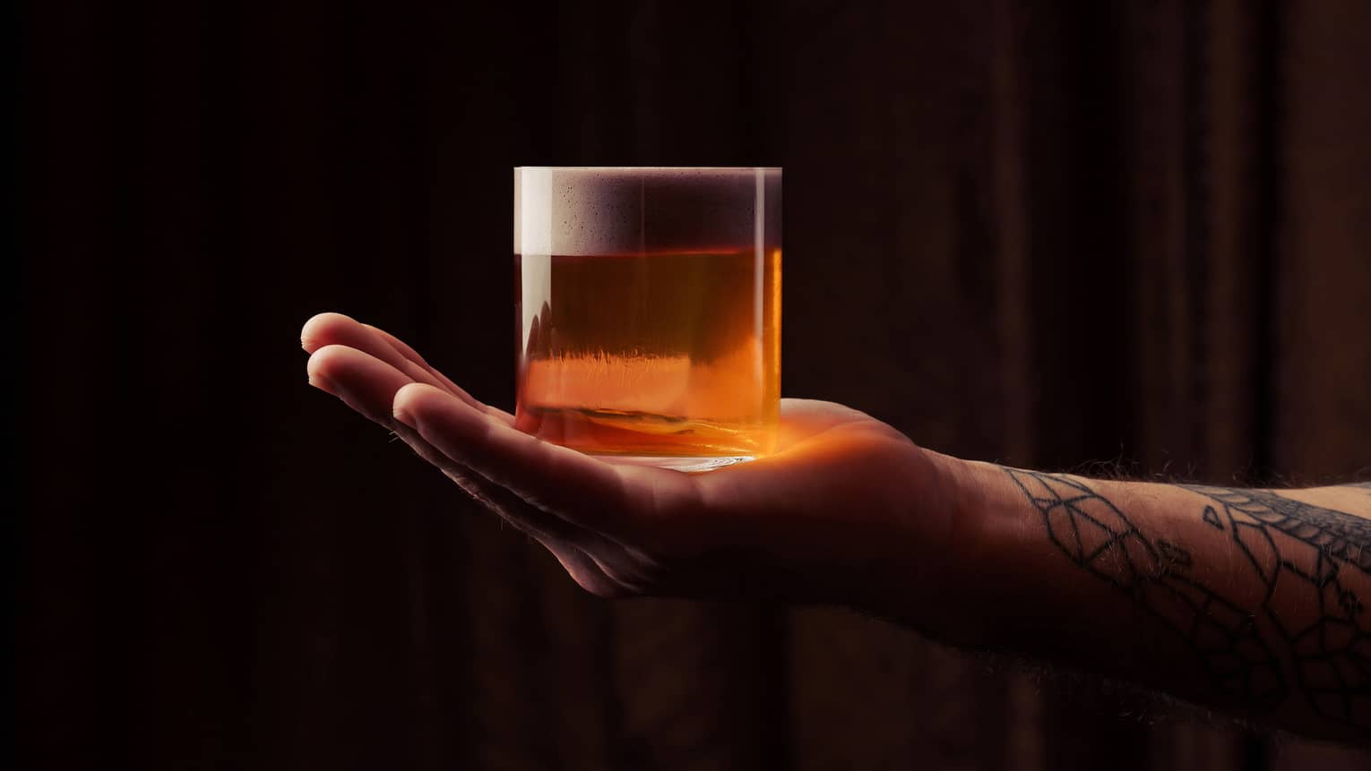 A tattooed arm reaches in from outside the frame and holds a rocks glass filled with an orange-colored cocktail on his outstretched hand