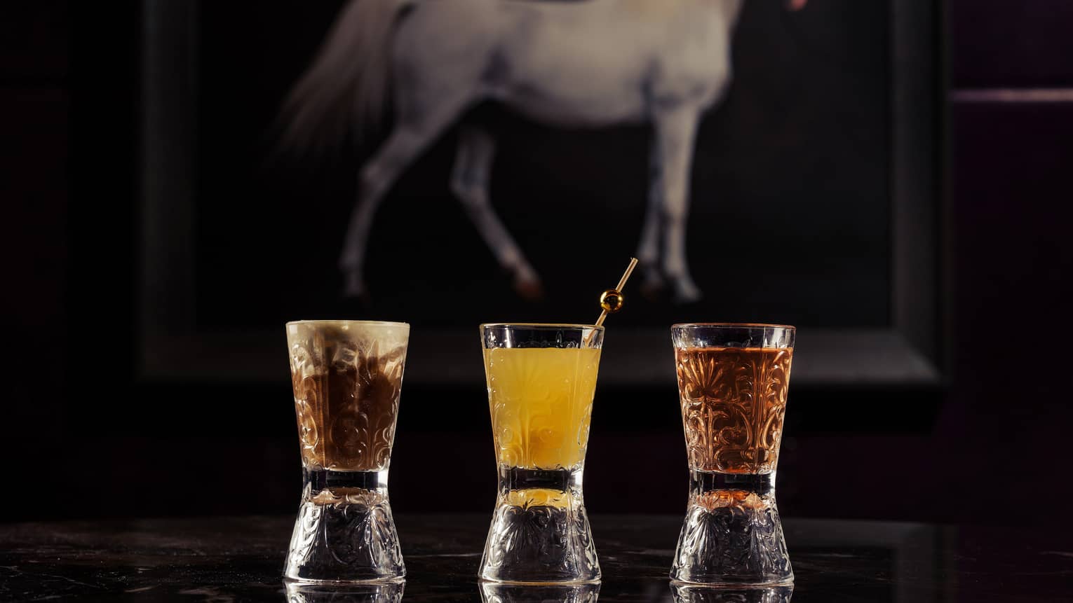 Three shot glasses in a row, the one on the far left holding a brown beverage, the one in the middle holding a yellow beverage and the one on the right holding a carmel-colored beverage, are placed on a dark table in front of a painting of a white horse