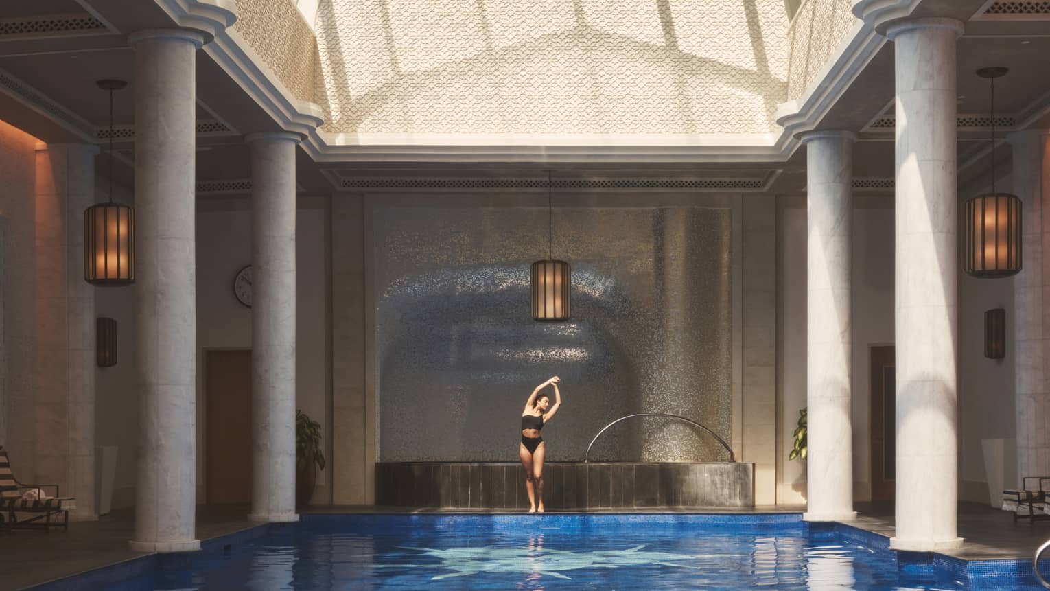 Person in swimsuit standing and stretching with arms overhead beside indoor pool flanked with white pillars under vaulted ceiling