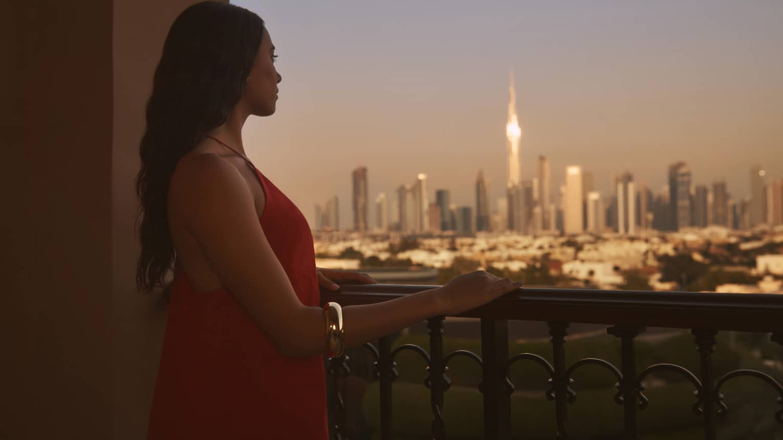 Person with long brown hair standing on balcony with hand resting on railing, looking out to a city at dusk