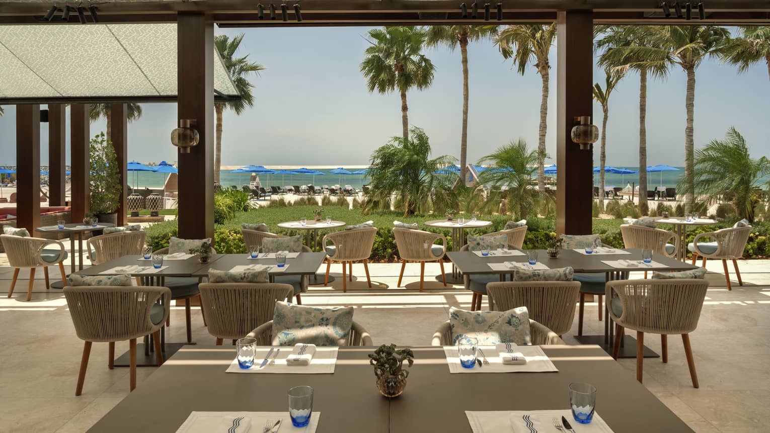 Restaurant terrace with multiple square tables opens up to a lawn lined with palm trees and the beach in the distance