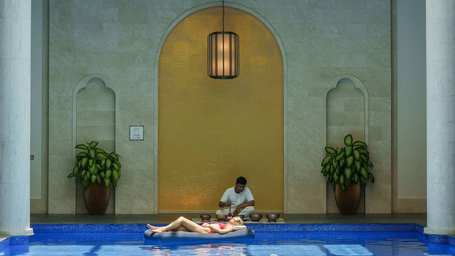 Person wearing red two-piece swimsuit lounges on a pool float in a pool framed by white columns as another person plays singing bowls on the pool deck