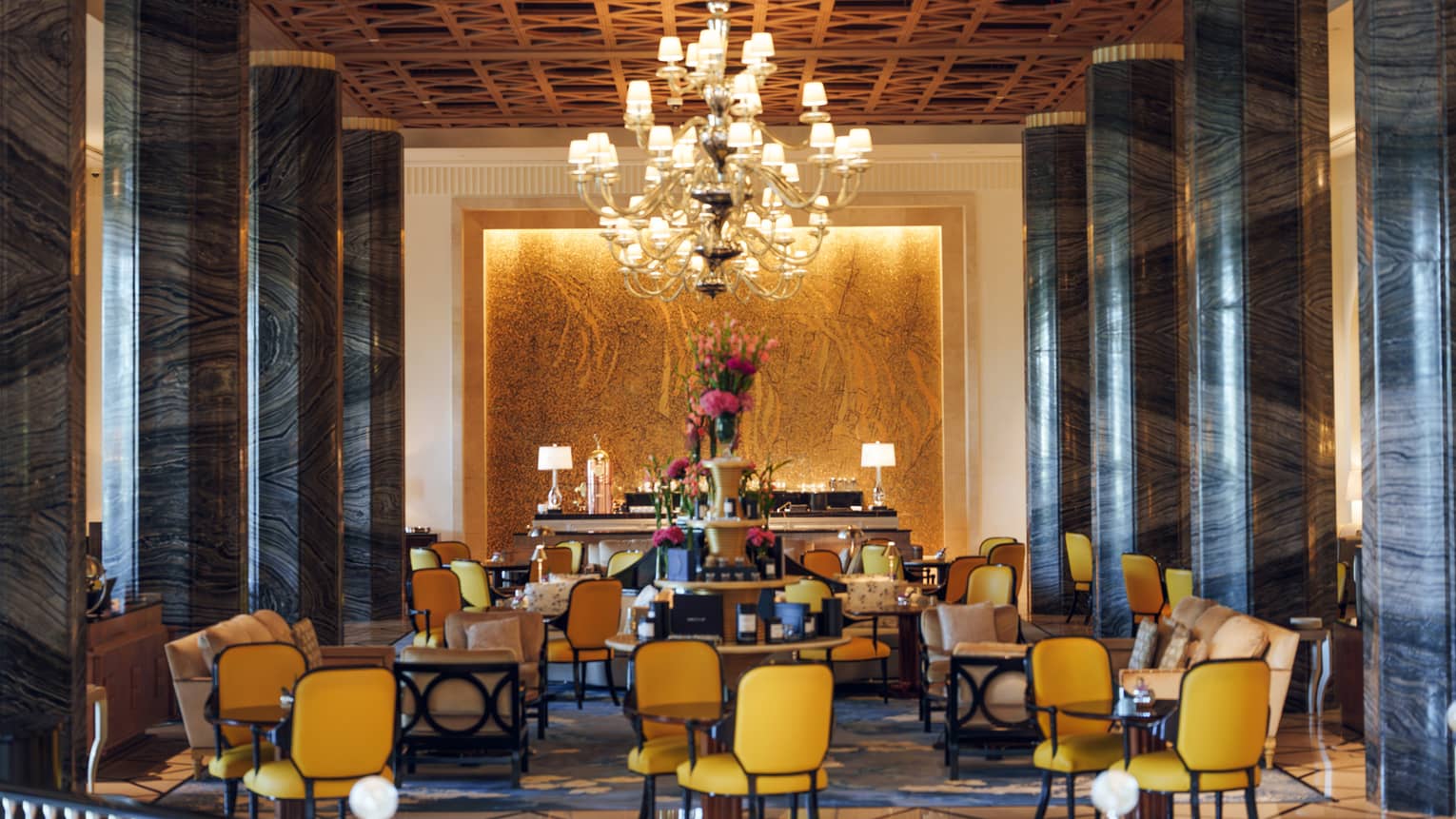 Hotel lounge furnished with dining tables, yellow chairs, a large central chandelier and two rows of tall black marble columns