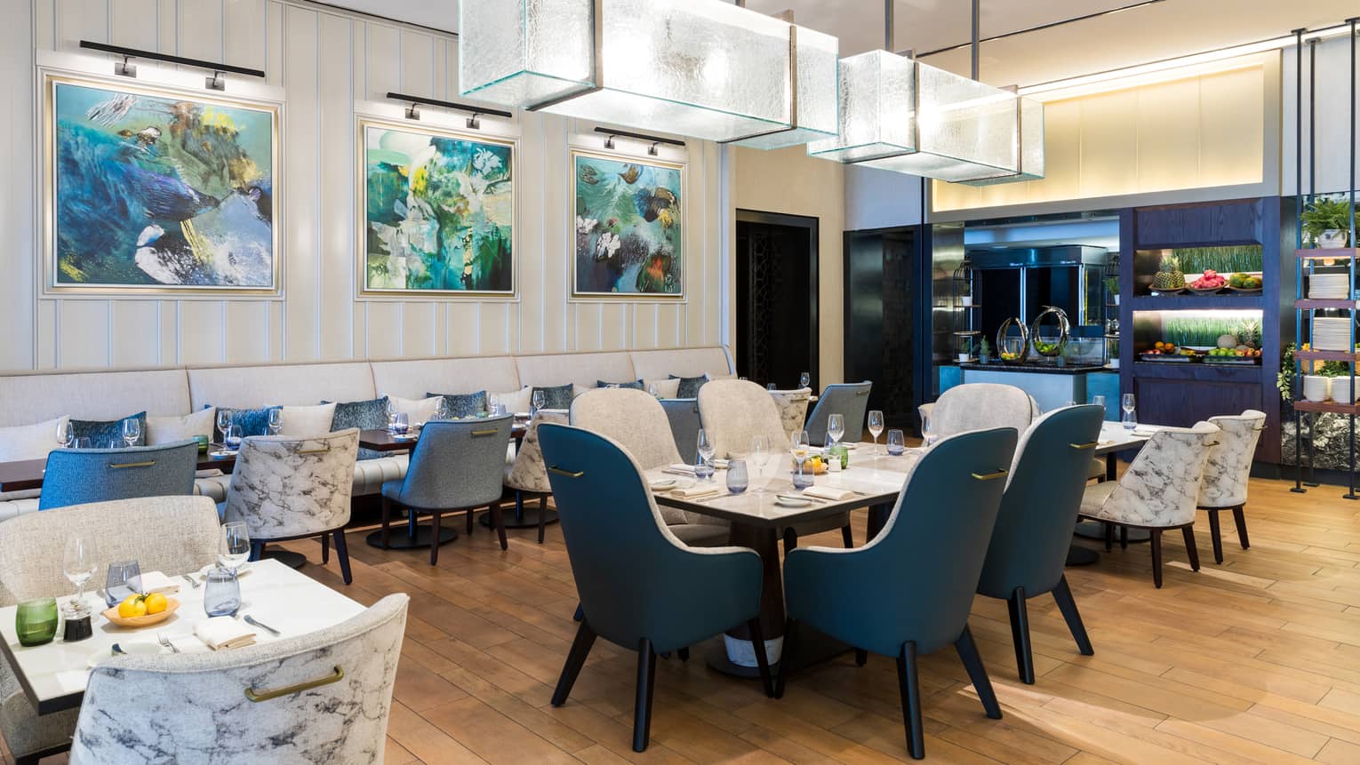 Interior of restaurant dining room with light-wood flooring, blue and white upholstered seating, large artwork on the walls and rectangular glass chandeliers hanging over the central tables